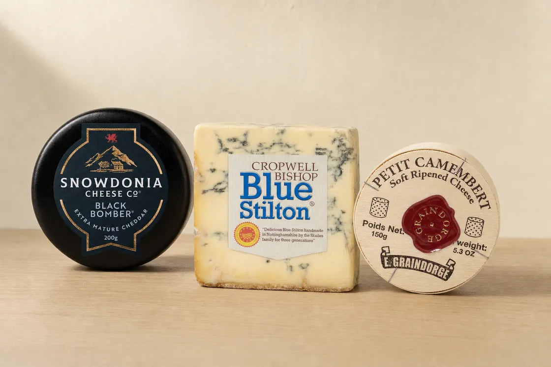 Three cheeses side by side with their labels facing out: a black waxed Snowdonia Black Bomber cheddar, a cut square of Cropwell Bishop Blue Stilton and a boxed E. Graindorge Petit Camembert.