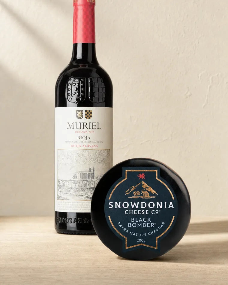 A bottle of Muriel Crianza Rioja, its label showing the estate building, standing beside a black waxed truckle of Snowdonia Black Bomber cheddar.